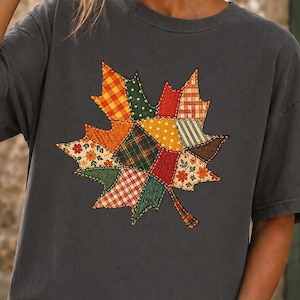 May include: A charcoal grey short-sleeve t-shirt featuring a large, central graphic of a maple leaf composed of various colourful patchwork patterns, including plaid, floral, and polka dot designs in shades of orange, green, red, and yellow.