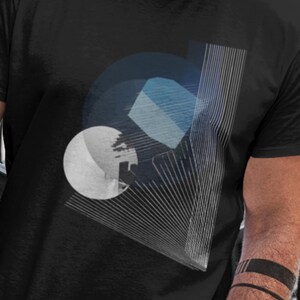 May include: Black t-shirt featuring an abstract geometric design in shades of blue, white, and gray. The design includes a circle, a polygon, and radiating lines. The shirt is short-sleeved and made of a soft material.