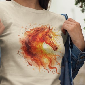 Könnte beinhalten: Ein cremefarbenes T-Shirt mit einer Aquarellillustration eines Pferdekopfes und einer fließenden Mähne in Orange-, Rot- und Gelbtönen. Das Design beinhaltet Farbspritzer-Effekte. Das T-Shirt wird getragen.