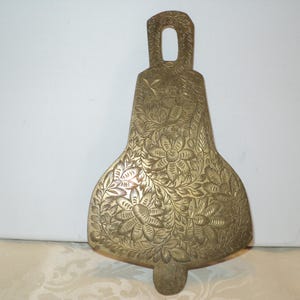May include: A brass-coloured, bell-shaped decorative item with a handle. The surface is intricately engraved with floral and leaf patterns. The bell has a rounded bottom and a small, rounded extension at the base.