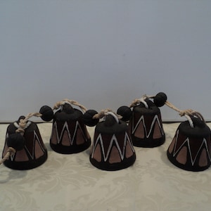 Puede incluir: Conjunto de cinco campanas decorativas. Cada campana es de color marrón oscuro con un diseño geométrico marrón claro y blanco. Están ensartadas con una cuerda de color natural y cuentas negras. Las campanas están dispuestas sobre una superficie estampada.