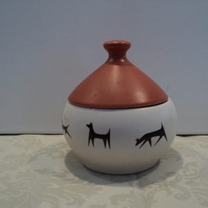 May include: A white ceramic jar with a terracotta-coloured lid. The jar features black silhouette designs of dogs. The jar is round with a lid that is cone-shaped. The jar is sitting on a patterned surface.