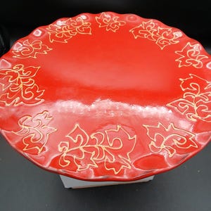 May include: A round, red ceramic plate with a scalloped edge. The plate is decorated with gold-colored floral designs. The plate is a decorative piece for serving food or displaying as a decorative item.