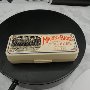 May include: A cream-colored plastic harmonica case with a rectangular shape. The lid features a black and white illustration of a band, the red text MARINE BAND, and the black text Made by M. HOHNER GERMANY No 1896 with decorative borders.