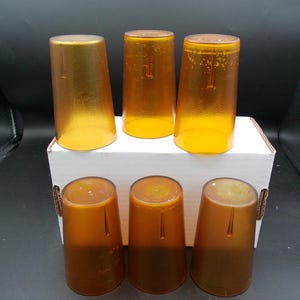 May include: Six amber-colored drinking glasses. Three glasses are upright on a white box, and three are below. The glasses have a textured surface and a tapered shape. The glasses are made of a translucent material.