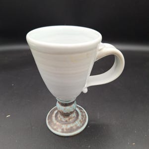 May include: A handmade white ceramic mug with a handle, set on a pedestal base. The base features a brown and turquoise color scheme. The mug has a conical shape and is suitable for hot or cold beverages.