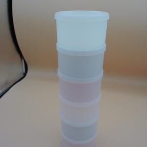 以下が含まれることがあります： 積み重ね可能な、複数のコンパートメントを備えた容器セット。セットには白い上部セクションがあり、ピンクとグレーの色合いのセクションが続きます。容器は円筒形で白い蓋が付いており、小さなアイテムの保管に最適です。