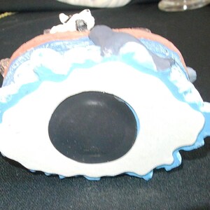 May include: A ceramic coin bank shaped like a seascape, featuring a white and blue design with a whale and a dog figurine. The bank has a large, black coin slot and is painted with various colors.