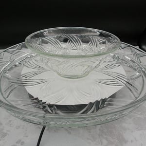 May include: A clear glass serving set consisting of a large, shallow circular platter and a smaller matching bowl. Both pieces feature a pressed glass design with radiating, curved, ribbon-like patterns that extend from the center outward.