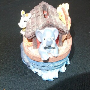 May include: A decorative figurine depicting Noah's Ark. The ark is a light brown color with a dark brown roof. A light blue elephant is in the front, with a white giraffe and cow on the sides. The base is blue, representing water.