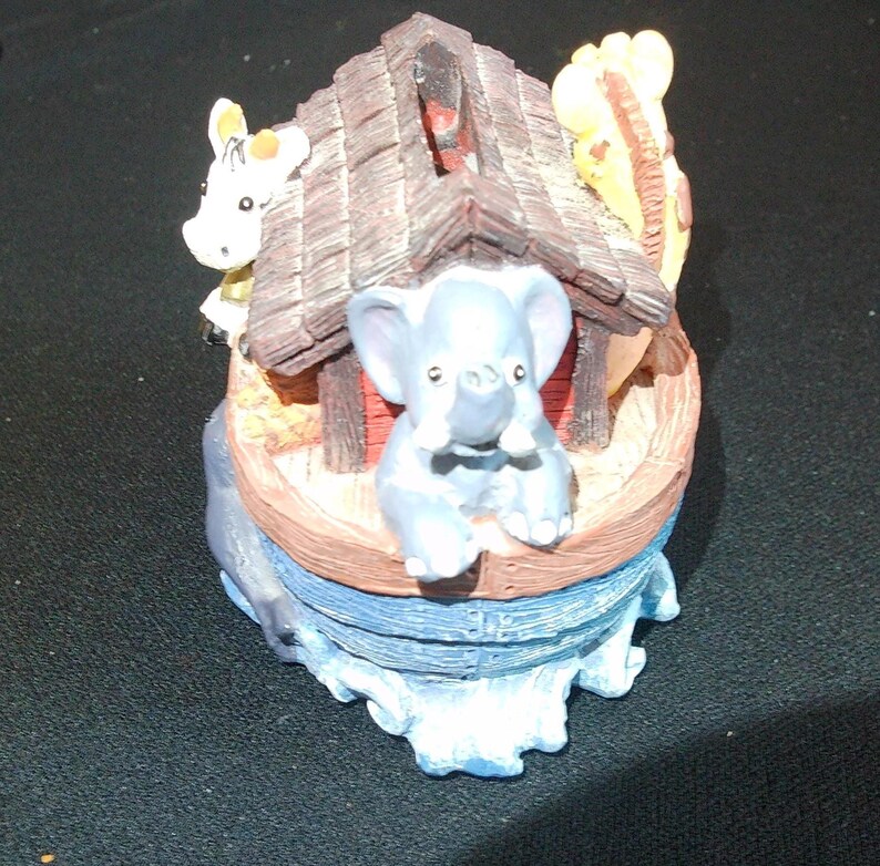 May include: A decorative figurine depicting Noah's Ark. The ark is a light brown color with a dark brown roof. A light blue elephant is in the front, with a white giraffe and cow on the sides. The base is blue, representing water.