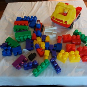 May include: A collection of colorful building blocks and a yellow and red toy truck with a small figure inside. The blocks are various sizes and colors, including blue, green, red, yellow, and purple. The toys are arranged on a white surface.