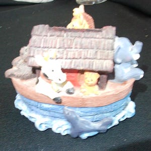 May include: A decorative figurine depicting Noah's Ark, featuring a boat with a brown roof and various animal figures, including an elephant, a giraffe, and a seal. The ark is surrounded by blue water, creating a whimsical scene.