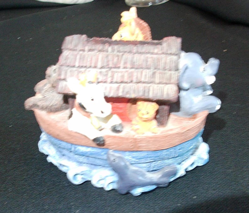 May include: A decorative figurine depicting Noah's Ark, featuring a boat with a brown roof and various animal figures, including an elephant, a giraffe, and a seal. The ark is surrounded by blue water, creating a whimsical scene.