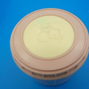 May include: A round, light pink container with a pale yellow lid. The lid features a raised, circular design with a honeycomb pattern in the center. The container sits against a bright blue background.