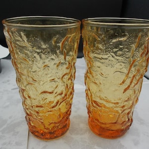 May include: Two amber-colored glass tumblers featuring a textured, organic surface pattern. The glass transitions from a lighter yellow hue at the top to a deeper, more saturated orange tone at the base, with a thick, uneven exterior finish.