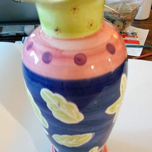 May include: A colorful ceramic vase with a ruffled rim. The vase features a light green neck with small yellow flower accents, a pink midsection with purple polka dots, and a blue base with white and green leaf designs. The base is multi-colored.