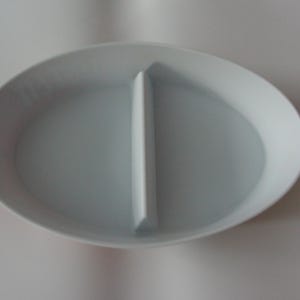 Puede incluir: Un plato de servicio blanco, ovalado y dividido. El plato está dividido por un divisor vertical, creando dos compartimentos separados. El plato tiene un acabado liso y brillante, adecuado para servir aperitivos o snacks.