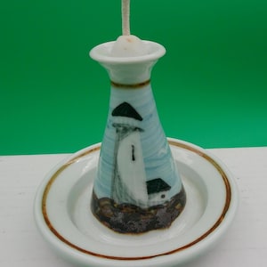 May include: A ceramic lighthouse-shaped candle holder with a white and blue design, featuring a lighthouse illustration. The candle holder sits on a white plate with a brown rim, and has a white wick.