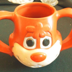 Puede incluir: Taza infantil de plástico naranja con dos asas grandes. La taza está decorada con una cara de zorro de dibujos animados, con ojos negros, hocico blanco y nariz roja. La taza está diseñada para un agarre fácil.