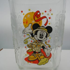 Puede incluir: Vaso de vidrio transparente con un diseño de Mickey Mouse en safari. La ilustración incluye un sol amarillo, una cinta roja y confeti. El vaso tiene forma cuadrada y detalles en relieve.