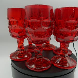 May include: A set of five red glass goblets featuring a faceted, geometric pattern on the bowl and a twisted stem design. Each goblet has a circular base and is crafted from translucent red glass, displaying a consistent texture throughout.