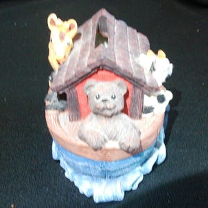 May include: A decorative figurine depicting Noah's Ark with a teddy bear, giraffe, and cow. The ark is brown with a red interior, and the animals are in various colors. The base is blue, representing water.