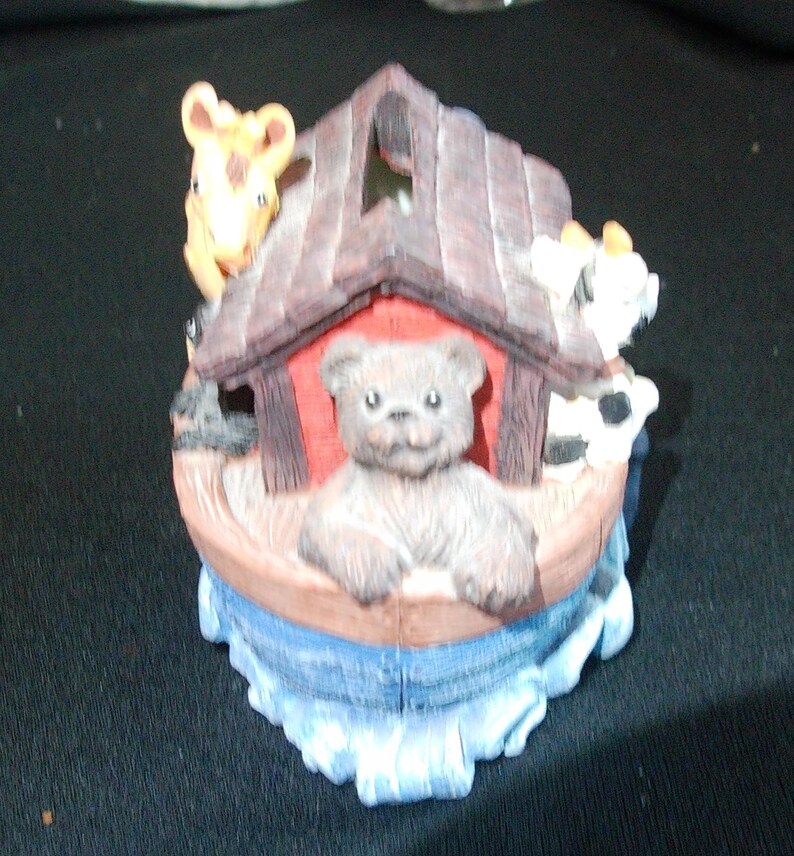 May include: A decorative figurine depicting Noah's Ark with a teddy bear, giraffe, and cow. The ark is brown with a red interior, and the animals are in various colors. The base is blue, representing water.