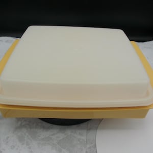 May include: A square yellow plastic container with a translucent white lid. The container features a flat base and a slightly raised rim, while the lid is smooth and opaque with a centered circular indentation. A black power cord extends from the side.
