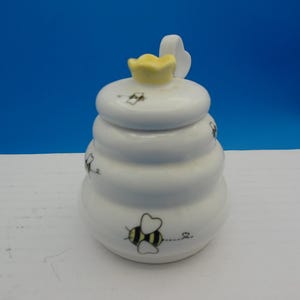 May include: White ceramic honey pot shaped like a beehive, with a yellow flower and white wing on the lid. The pot is decorated with black and yellow bee illustrations.