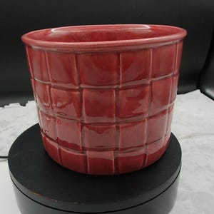 May include: A cylindrical ceramic planter with a glossy, deep red finish. The exterior surface features a raised, grid-like pattern of square tiles that creates a textured, woven appearance around the entire circumference of the vessel.