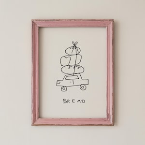 May include: A framed artwork with a pink, distressed wooden frame. The artwork features a simple black line drawing of a car carrying a stack of bread on its roof, with the word "BREAD" below.