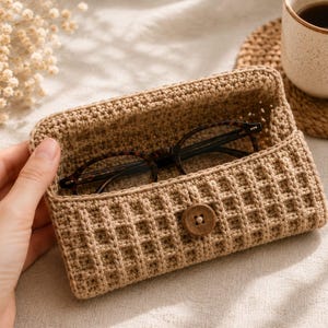 May include: A beige crochet glasses case featuring a waffle-like textured pattern and a single wooden button closure. The rectangular pouch is open, revealing a pair of dark brown tortoiseshell-patterned eyeglasses resting inside the structured compartment.