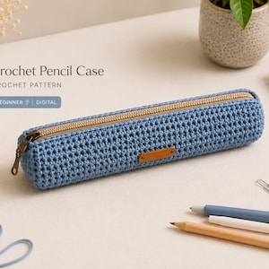Può includere: Un astuccio blu all'uncinetto con cerniera beige e una piccola etichetta in pelle marrone. L'astuccio è cilindrico e si trova su una superficie chiara, con matite, graffette e un taccuino nelle vicinanze. Il testo "Crochet Pencil Case" è visibile.