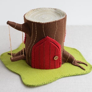 May include: A felt tree house with a red door, a brown trunk, and a green base. The tree house has a swing attached to the side.