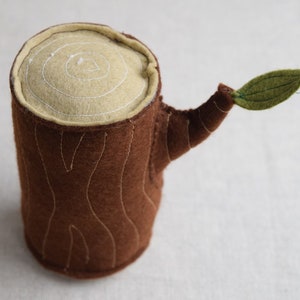 May include: A brown felt tree stump with a light brown felt top and a green felt leaf. The stump has white stitching that resembles tree bark.