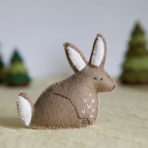 May include: A brown felt bunny with white felt ears and a tail. The bunny is sitting on a light brown surface with two green felt trees in the background.