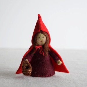 Puede incluir: Una muñeca de fieltro con una capa roja con capucha y un vestido burdeos. La muñeca lleva una cesta marrón.
