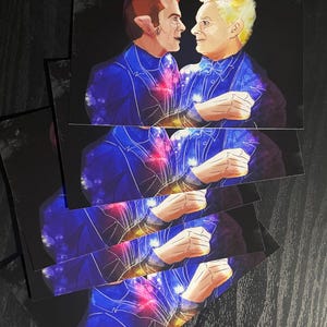 May include: A stack of rectangular art prints featuring a digital illustration of two figures facing each other. The figures are dressed in dark blue suits with a vibrant galaxy pattern of purple, blue, and red nebula clouds across their clothing.