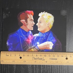 May include: A rectangular art print featuring a digital illustration of two figures facing each other, set against a dark background. The figures wear blue suits with galaxy patterns. A wooden ruler is placed below, showing a width of 20 cm.