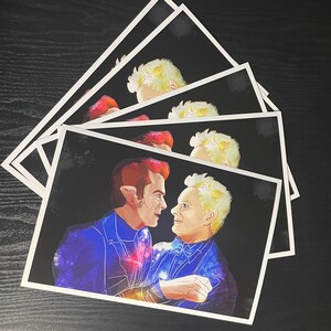 May include: A stack of rectangular art prints featuring a digital illustration of two figures facing each other against a black background. The figures have stylised, galaxy-patterned clothing in shades of blue, purple, and orange, with light hair.