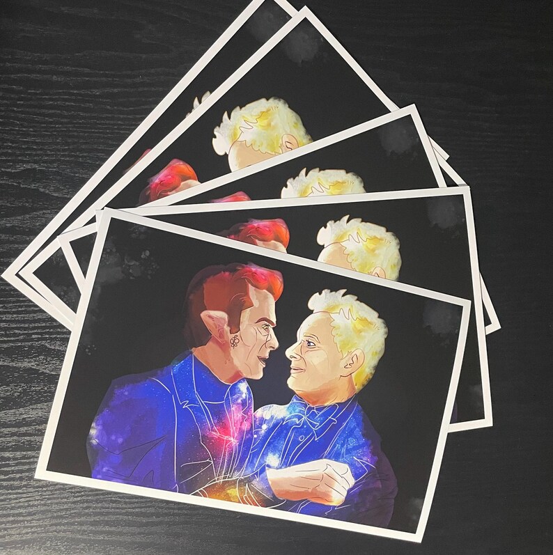 May include: A stack of rectangular art prints featuring a digital illustration of two figures facing each other against a black background. The figures have stylised, galaxy-patterned clothing in shades of blue, purple, and orange, with light hair.