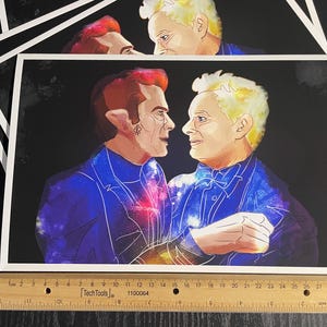 May include: A rectangular art print featuring a digital illustration of two figures facing each other, set against a dark background. The figures wear blue suits with galaxy patterns. A wooden ruler measuring 30 cm is placed along the bottom edge.