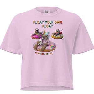 Può includere: T-shirt rosa chiaro con la scritta "FLOAT YOUR OWN FLOAT" e "BEWITCHED CUTIES". La grafica presenta bradipi in anelli gonfiabili, uno con un corno di unicorno. La maglietta è realizzata in tessuto morbido.