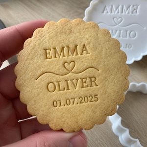 Op de afbeelding: Een rond koekje met een geschulpte rand voorzien van een reliëf met de namen EMMA en OLIVER, een klein hartje en de datum 01.07.2025. Een witte plastic koekjesstempel met hetzelfde ontwerp is zichtbaar op de achtergrond.