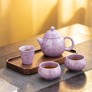 May include: A lavender ceramic tea set with floral accents. The set includes a teapot, two teacups, and a small cup, all arranged on a wooden tray. The tea cups and teapot are decorated with delicate pink flowers.