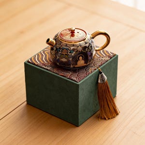Puede incluir: Una tetera decorativa con asa y boquilla doradas, colocada sobre una caja verde con tela estampada. La tetera presenta intrincados diseños en negro, dorado y azul, con una tapa dorada. Una borla marrón cuelga de la caja.