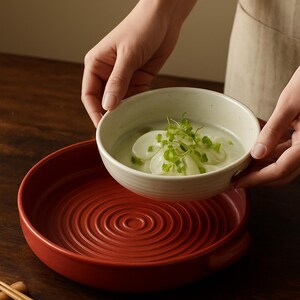 May include: A person holding a white ceramic bowl filled with soup and garnishes above a red ceramic serving dish. The serving dish has a concentric circle design. Chopsticks rest on a dark wooden surface.
