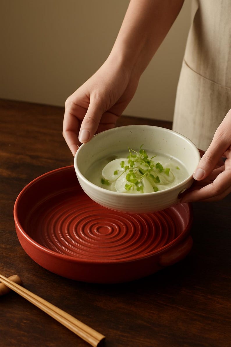 May include: A person holding a white ceramic bowl filled with soup and garnishes above a red ceramic serving dish. The serving dish has a concentric circle design. Chopsticks rest on a dark wooden surface.