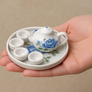 Puede incluir: Miniatura de juego de té de porcelana blanca con diseño floral de rosas azules. El juego incluye una tetera, cuatro tazas pequeñas y una bandeja. La tetera tiene tapa y asa.